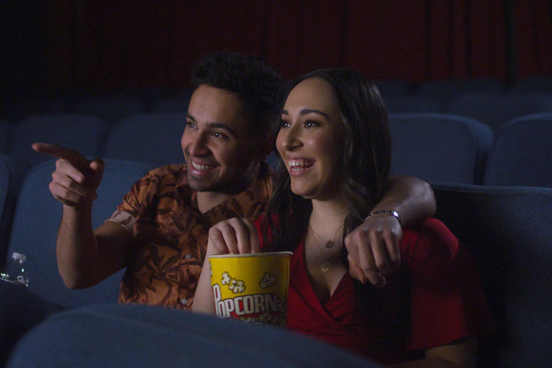 A man and a woman eating popcorn and watching a movie in a theatre. The seats are dark blue, set up in multiple rows.