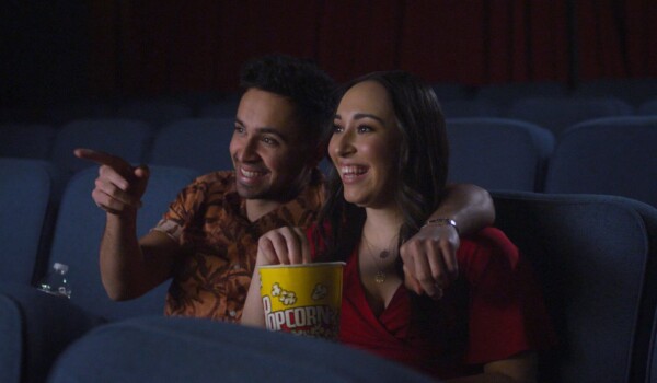A man and a woman eating popcorn and watching a movie in a theatre. The seats are dark blue, set up in multiple rows.