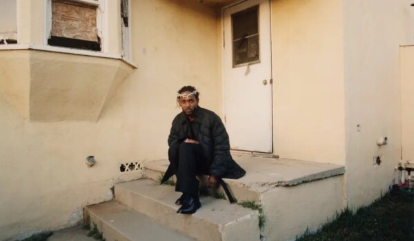 Kendrick Lamar, wearing a crown of thorns, sits on the steps outside a house